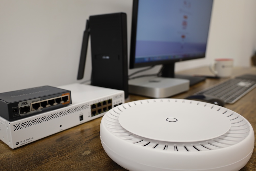 MikroTik hEX S and CAP ax Home Network Setup Example MikroTik hEX S router, 8-port PoE switch, and CAP ax Wi-Fi 6 access point on a desk beside a computer, showing a typical home network setup.